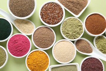 Different superfood powders in bowls and spoons on pale green background, flat lay