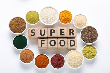 Different superfood powders in bowls and words made of wooden cubes on white background, flat lay