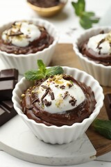 Tasty pudding with whipped cream in bowls and pieces of chocolate on white wooden table, closeup