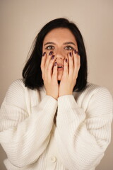 A young woman holds her hands over her face, her eyes wide open and her mouth open in genuine surprise. The interesting facial expression conveys a moment of surprise. The focus is on her emotions.