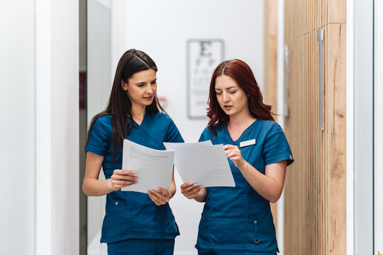 Two female doctors in blue scrubs are standing in a modern hospital corridor, carefully reviewing patient medical records and discussing treatment strategies