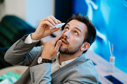 Businessman sitting at his desk applying eye drops to relieve dry eyes or conjunctivitis, a common problem for office workers spending long hours in front of computer screens