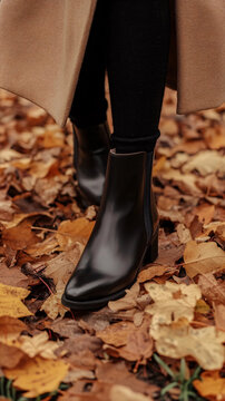 A person is wearing black leather ankle boots while walking among autumn leaves on the ground.