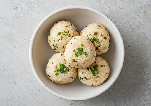 A top view of five rice balls with green herbs in a white bowl on a gray surface with natural light