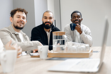 Multinational group of diverse professionals engaged in collaborative meeting at office table, sharing ideas and discussing business strategies while enjoying coffee.