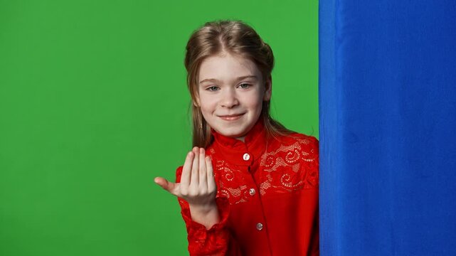 Smiling girl child peeks out waves hand and shows come here gesture, positive expression, isolated on chroma key green screen studio background mockup.