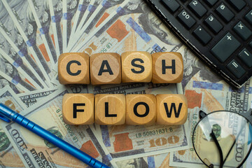 Wooden block with "CASHFLOW" written on it arranged on top of several $100 bills, surrounded by a pair of glasses, a black calculator and a blue pen. Business and finance concept