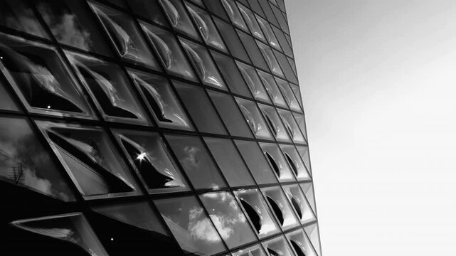 Black and white modern glass facade reflecting clouds, angled view of building exterior, smooth curved surfaces creating abstract patterns, light and shadow contrast, minimalist design