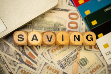 A photo depicting saving, with some wooden blocks with the word "SAVING" written on them surrounded by some dollar bills, debit or credit cards, and a laptop. Investment concept