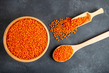 Red lentil grains in a wooden bowl and spoon on a gray background. Top view of dried orange lentils inside the bow