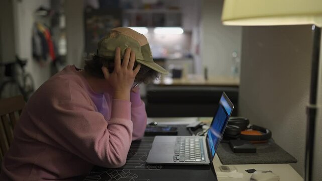 Tired female student in a pink sweatshirt watches an online lecture late at night in a small, dimly lit apartment. She looks bored and exhausted during a tedious online class or video call. 