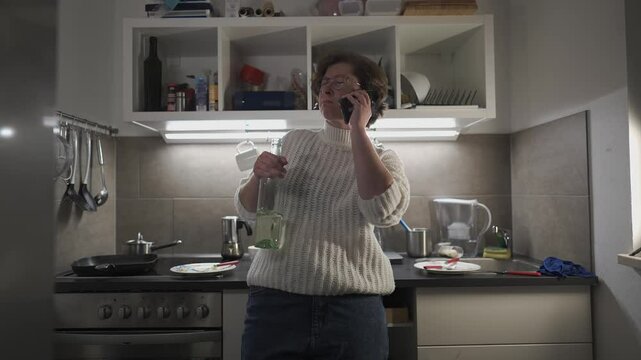 A lonely, drunk Caucasian woman in her mid-30s stands in a small kitchen, wearing glasses, jeans, and a white sweater, drinking white wine from a bottle and speaking on the phone in visible despair