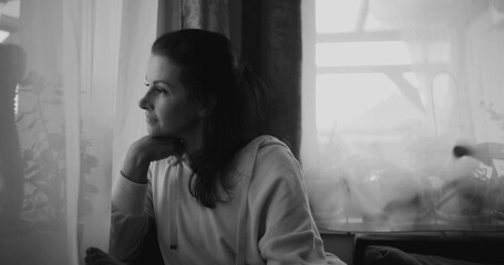 Woman gazing out window, resting chin on hand in thoughtful pose, sitting in peaceful home setting surrounded by plants and curtains, reflecting in monochromatic tones