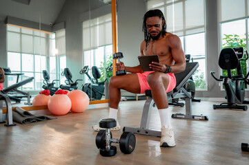 A shirtless man sits on a bench in the gym holding a dumbbell and looking at a tablet, combining strength training with guided digital instruction.