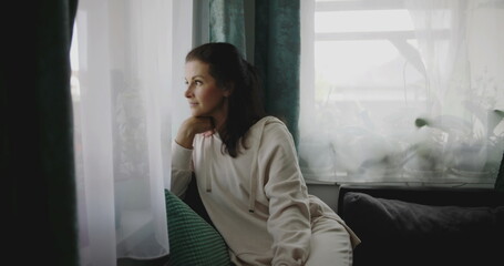 Fototapeta premium Woman leaning forward on sofa, gazing out window with chin resting on hand, surrounded by plants and curtains in a quiet home environment, reflecting with calm demeanor