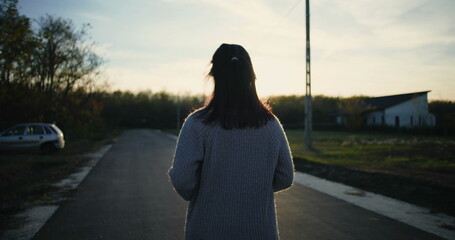 Woman walking down a quiet road at sunset, back turned, peaceful countryside atmosphere, car parked to the side, serene outdoor setting, rural surroundings captured