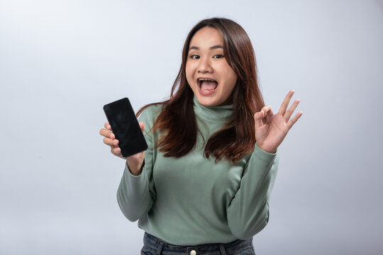 Smiling Asian woman in green sweater holding smartphone with blank screen and making OK hand gesture, winking playfully against plain gray background. Confident approval and tech lifestyle concept