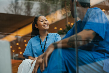Happy female healthcare professional talking with male coworker while sitting in hospital building seen through glass