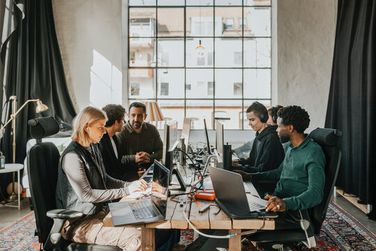 Group of multiracial computer professionals working on desktop PC while sitting at desk in tech startup office