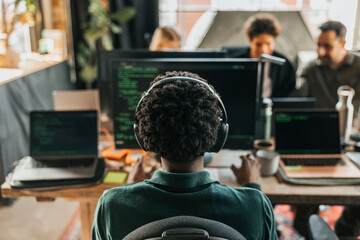 Rear view of curly haired male computer programmer wearing wireless headphones coding on desktop PC while sitting on cha