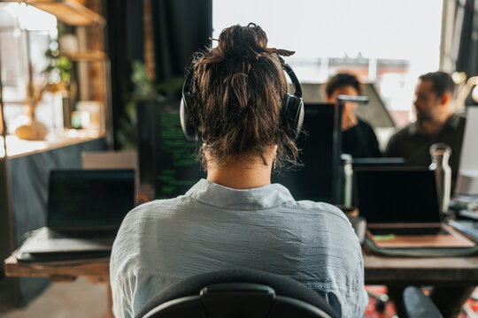 Rear view of female IT professional wearing wireless headphones working on desktop PC in office