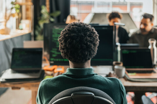 Rear view of curly haired male computer programmer coding on desktop PC while sitting on chair in tech startup office
