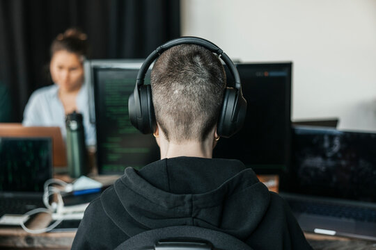 Rear view of non-binary IT professional wearing wireless headphones coding on desktop PC while working in office