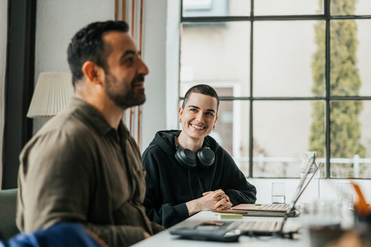 Happy non-binary software engineer sitting with male colleague at desk in tech start-up company