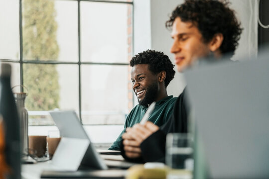 Happy young male software engineer sitting with colleague at desk in tech startup office