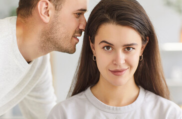 Obraz premium Close up portrait of handsome young man whispering something in the ear of smiling brunette confident woman telling her secret or gossip sharing news. Young couple talking indoors.