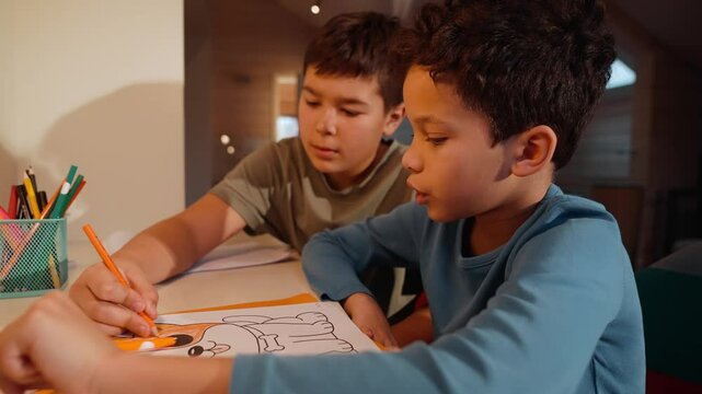 Two school-aged Latino boys concentrate on drawing together at a white desk. One boy attentively colors a picture with an orange pencil, while the other leans in closely, helping him to draw.