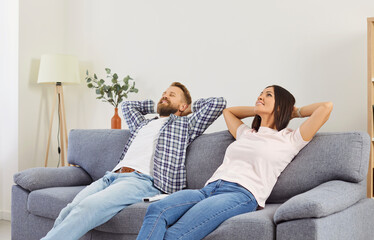 Smiling young family couple relaxing while sitting with hands behind their heads on couch in living...