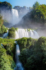 Marmore Waterfalls cascade down lush, green cliffs enveloped in mist, creating a breathtaking natural spectacle. The serene landscape showcases the beauty of Italy's renowned waterfall