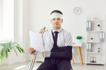 Portrait of a young handicapped injured man with broken arm and bandaged head after accident working in office holding document in hands and looking at camera. Insurance and compensation concept.