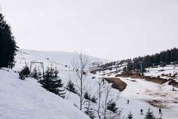 Partially snow-covered ski slope with ski lift under a cloudy sky.