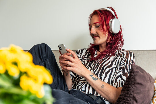 Non-binary person relaxing on sofa using smartphone and listening music with headphones