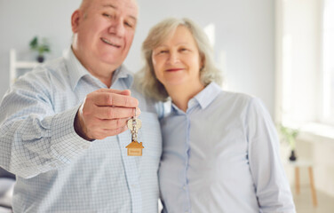 Happy senior couple holding a key in hands standing in the living room at home looking at camera enjoying real estate purchase, smiling and celebrating moving day. Relocating, mortgage concept.