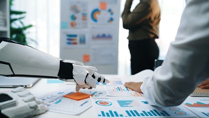 A robot arm collaborates with humans in a modern office, analyzing data charts and graphs to drive strategic decisions and innovative business solutions.Noogenesis