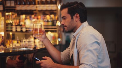 Smart man drinking cocktail in Old fashioned waiting friends or couple while chatting on smartphone to wait at counter bar with bartender preparing drinks at nightclub Friday meeting night. Vinosity.