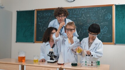 Boy pour solution in beaker while american girl inspect colored liquid. Energetic student support his friend doiing experiment while teacher looking children explore and discover innovation. Pedagogy.