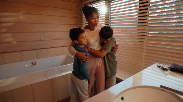 In the morning, a happy Black mother in her early 30s hugs her two dark-haired school-aged sons, stroking their heads, talking to them, while both fool around with her and laugh in the bathroom.
