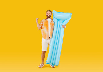 Funny young smiling man wearing beach clothes in sunglasses holding inflatable mattress isolated on studio yellow background. Happy tourist is going on summer holiday trip. Vacation concept.