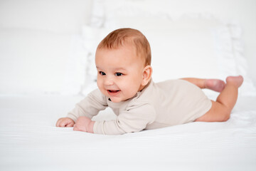 a baby, a smiling baby on a white bed on his tummy at home, a healthy, joyful little baby boy in a...