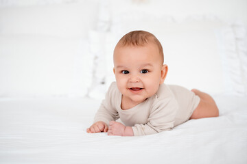 a baby, a smiling baby on a white bed on his tummy at home, a healthy, joyful little baby boy in a...