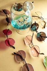 Summer composition with retro-style sunglasses in different colors around a blue cocktail with ice and lemon slices, on a beige background.