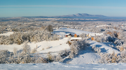 Farm on hill in winter landscape, forest covered with snow, mountain in distance