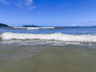 A tranquil view of the sea on a sunny day where the gentle waves break on the sand. Clear blue skies complement the serene scenery, with small islands in the background reflecting in the water.