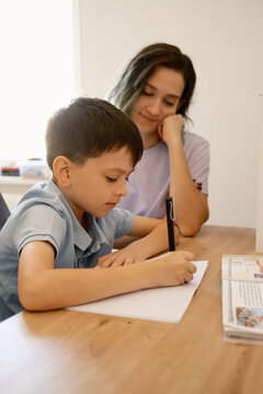 A mother guides her son with his homework, creating a supportive learning environment. The mom watches attentively as the boy focuses on writing in a notebook.