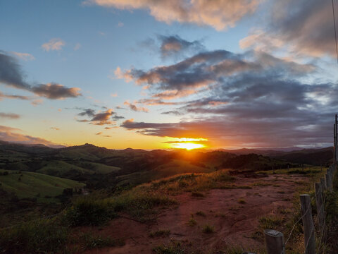 A stunning sunset over a backdrop of green hills, with the soft afternoon light illuminating the cloud-filled sky. The wooden fence and the road on the edge of the countryside add depth.