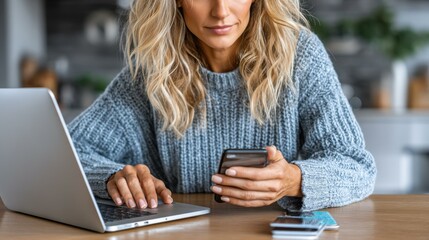 Woman in blue sweater types on laptop while holding cellphone indoors with credit cards on the table surface